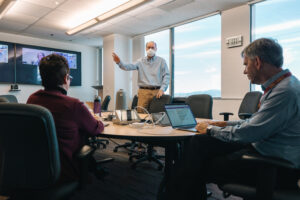 Office collaboration session during hybrid team meeting with workers seating around round table engaged in discussion with virtual attendees on monitor screen