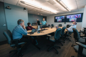 Office collaboration session during hybrid team meeting with workers seating around round table engaged in discussion with virtual attendees on monitor screen