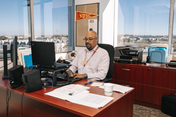 Person wearing button down shirt sitting by corner desk typing on computer laptop in front of windows overlooking the city